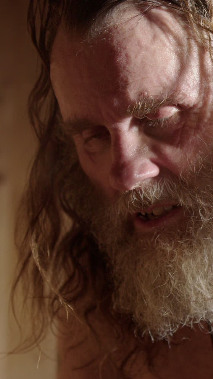 Scruffy man pours water on hot stones steaming up wooden sauna. Vertical