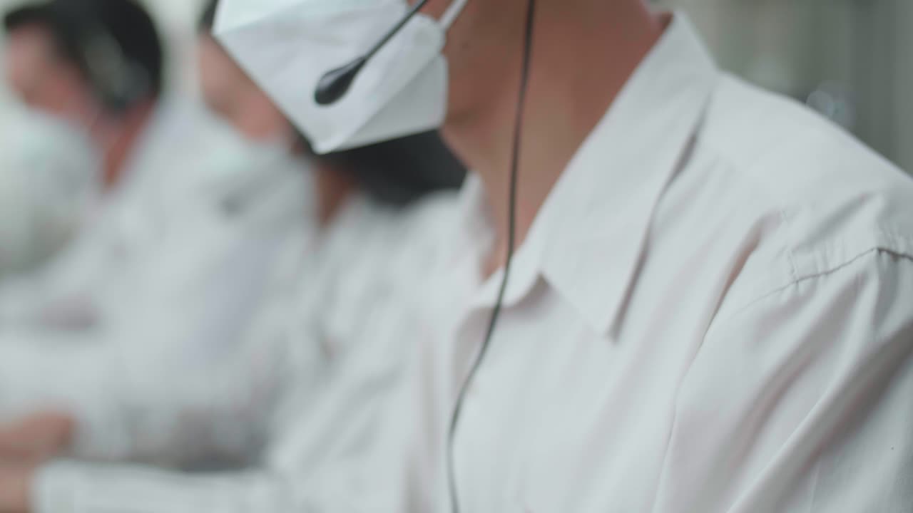 close up de un hombre asiático agente de centro de llamadas con auriculares y máscara escribiendo en la computadora mientras habla con el cliente en la llamada durante el trabajo con colegas en la oficina