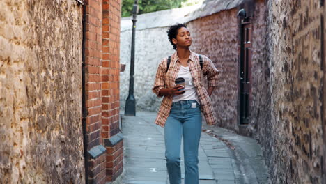 young woman wearing a plaid shirt and blue jeans walking towards camera in an alley between old stone walls holding a takeaway coffee, selective focus