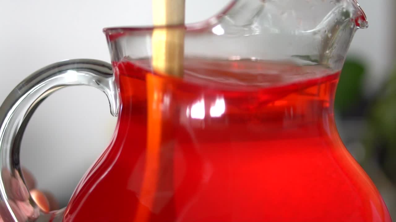Stirring red drink mix in glass pitcher of water with wooden spoon