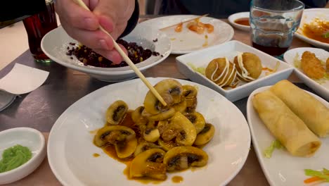 We-see-Asian-dishes-of-mushroom-with-soy-another-of-octopus-balls-black-rice-and-Vietnamese-rolls-a-hand-appears-with-chopsticks-picking-up-a-mushroom-there-is-wasavi-a-glass-and-a-bottle