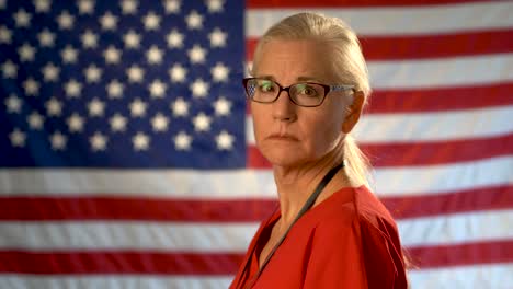 Medium-tight-portrait-of-the-back-of-blonde-nurses-head-as-she-turns-and-looks-at-camera-looking-happy-wearing-glasses-with-out-of-focus-US-flag