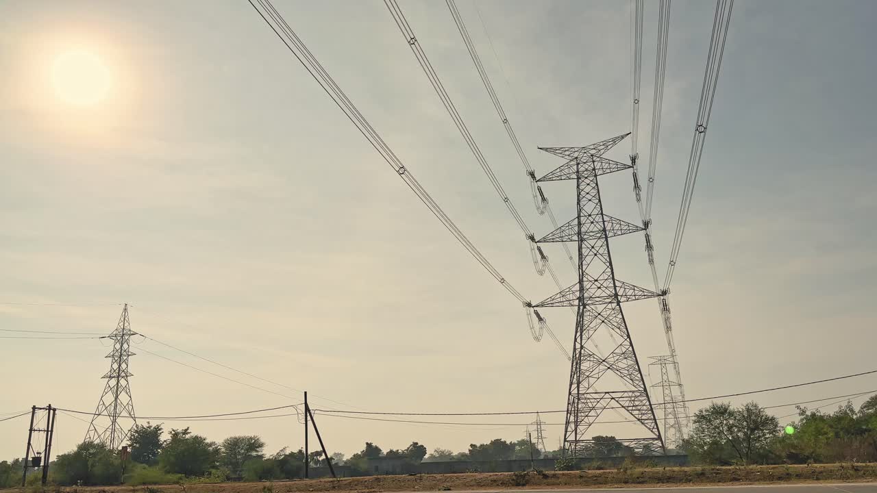 Wide view of steel transmission towers carrying high-voltage power lines across open land, sunlight filtering through hazy sky with trees and utility poles on horizon