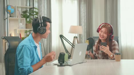 young asian woman holding phone and wearing headphones singing into microphone while recording podcast with male host in studio