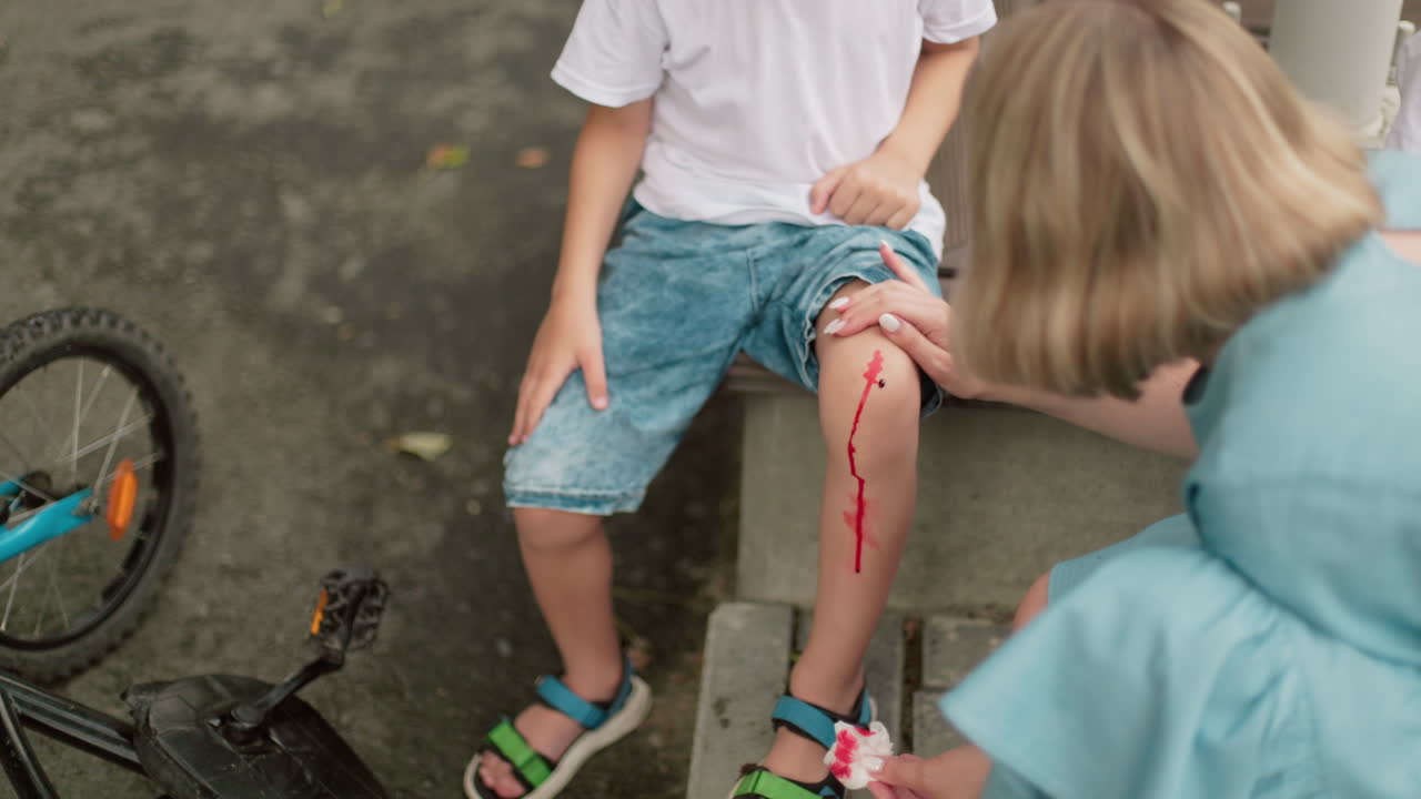 A woman carefully attends to a child's wounded knee using a blood-stained tissue, while the child sits nearby with a fallen bicycle on the ground