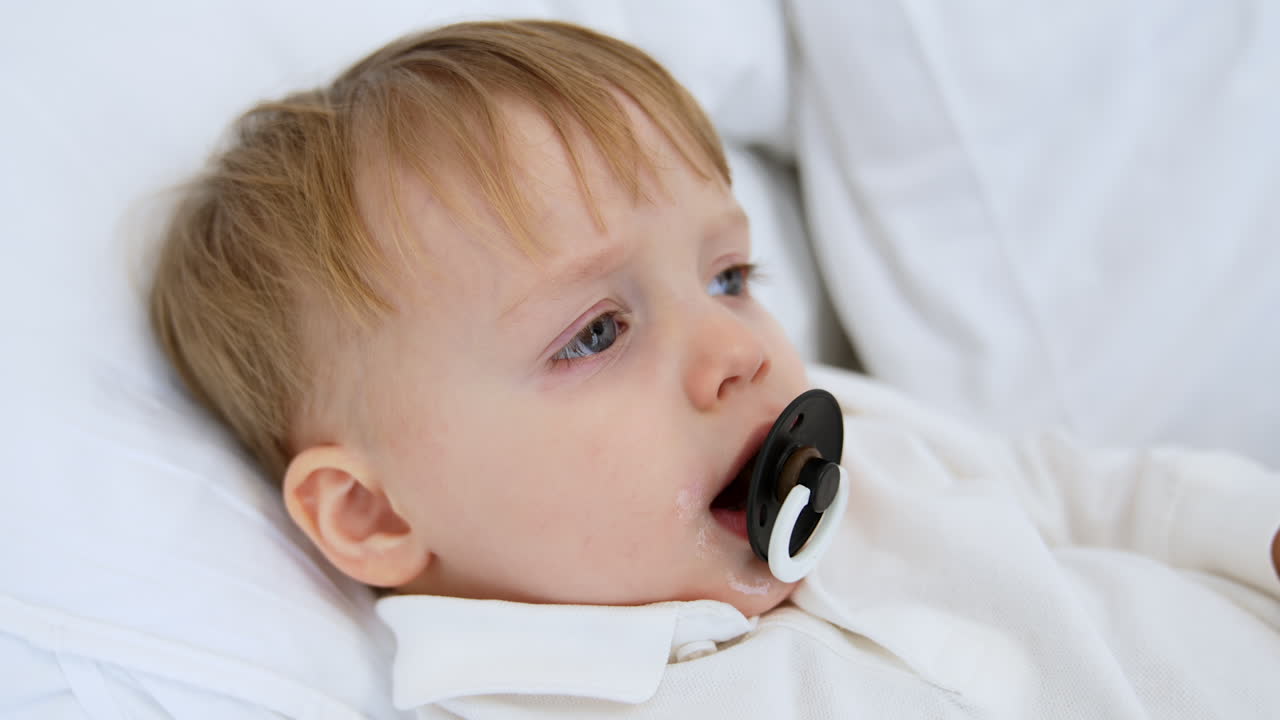 Caucasian toddler sucking a pacifier lies on the bed. Side view close up portrait of baby falling asleep.
