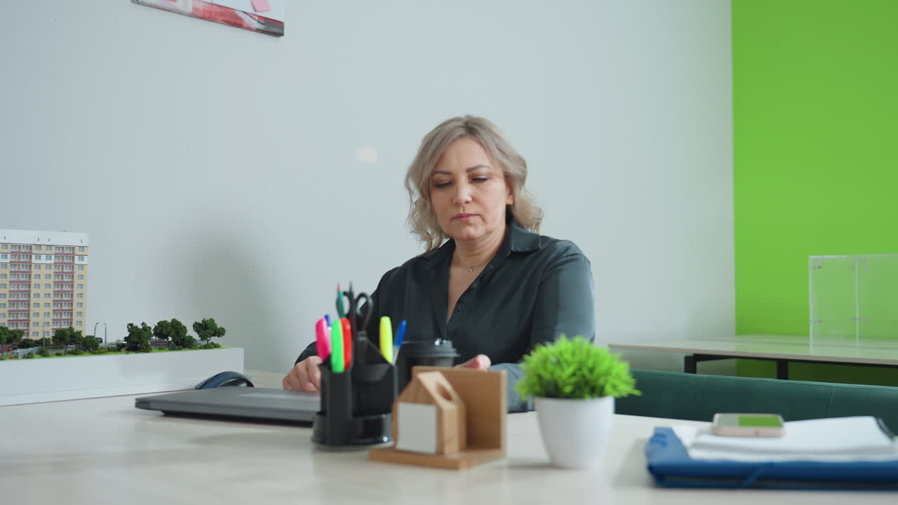 Realtor takes seat in modern office, preparing for day work beside building model, with coffee on table and workspace filled with essential supplies