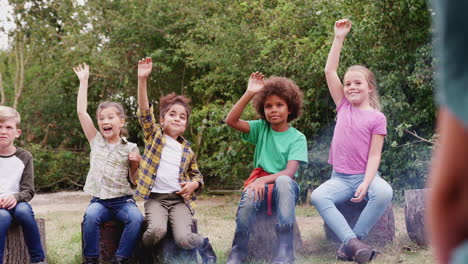 group of children on outdoor activity camping trip sitting around camp fire with arms raised answering question