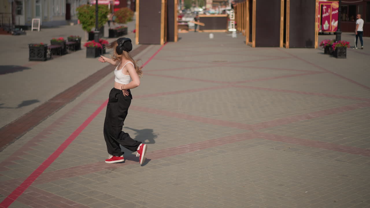 Ballerino di strada solitario che prova urban groove, pantaloni larghi e sneakers rosse, piazza illuminata dal sole con vetrine, passi ritmici sicuri, abbigliamento casual, prova di coreografia, ampio scatto cinematografico