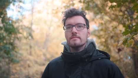 joven europeo con gafas y barba de pie en un bosque de otoño mixto, mirando a la distancia