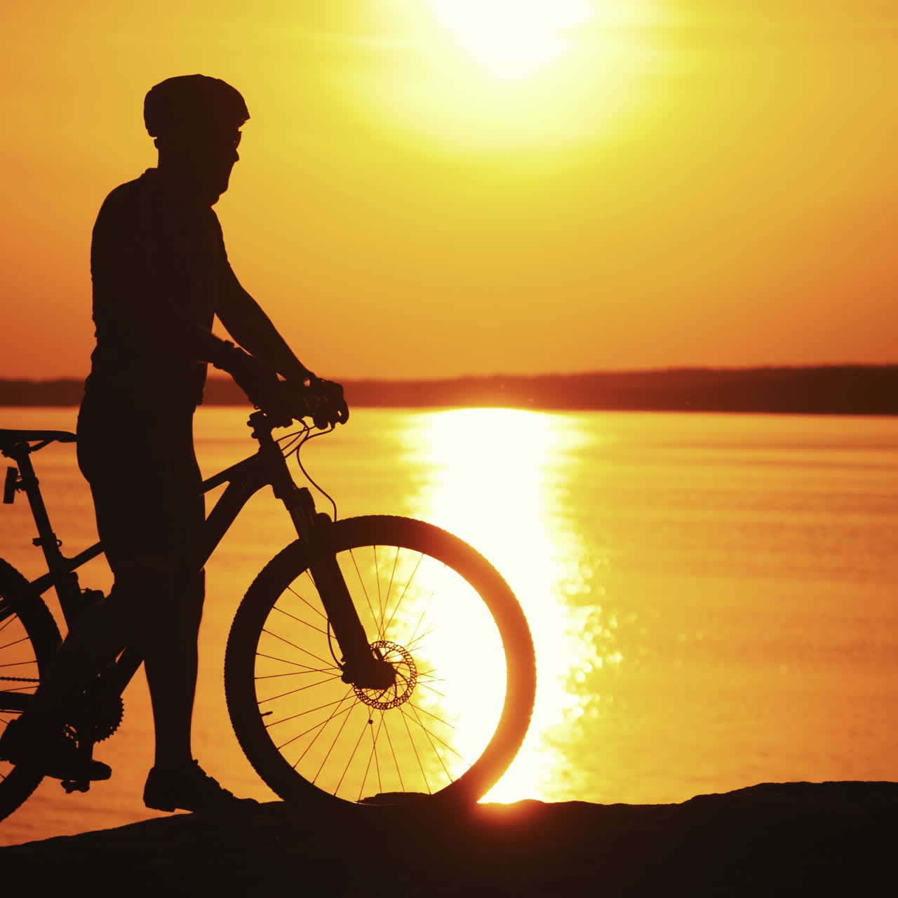 Silhouette of a cyclist near the river at sunset. Sporting man in helmet and glasses rides his bike on the background of beautiful sunset over the water surface.