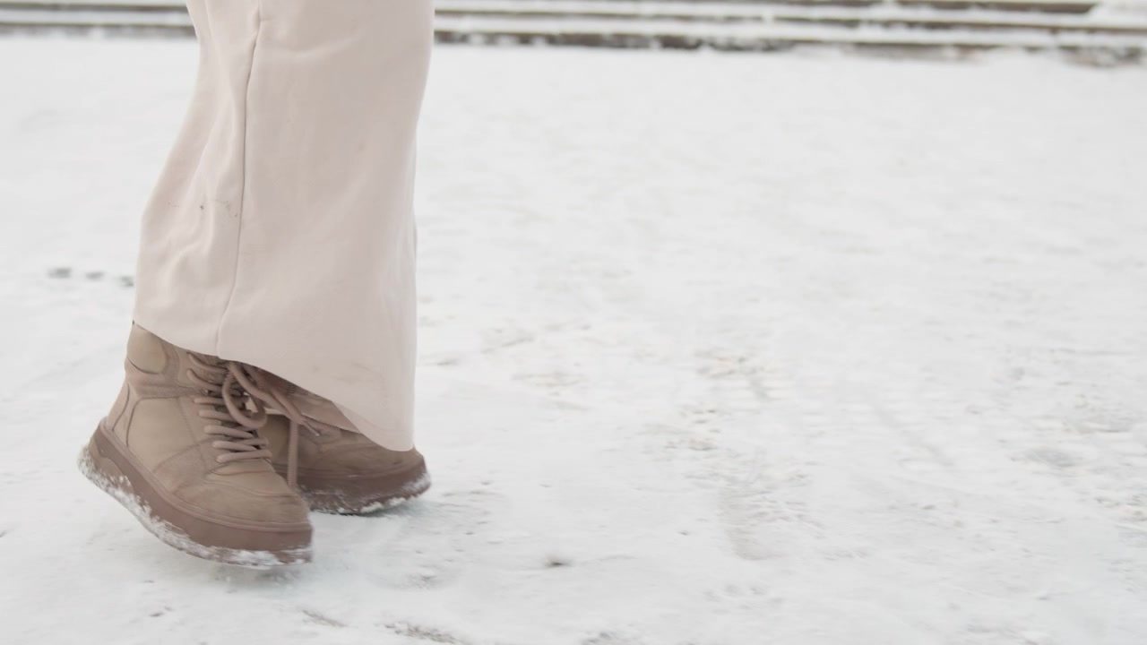 Vista de cintura para abajo de una persona caminando por un pavimento urbano cubierto de nieve con pantalones anchos beige y botines marrones con cordones, mostrando el talón levantado, luz invernal suave y un ambiente callejero frío de temporada.
