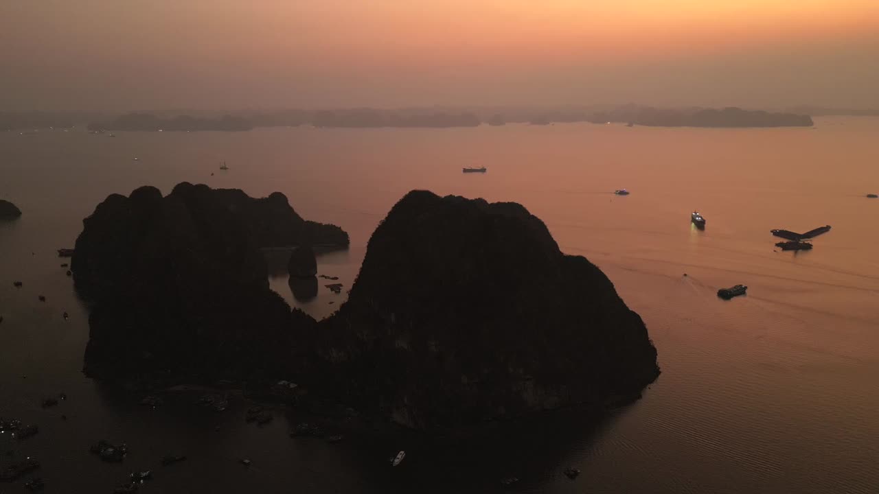 Aerial view of dramatic limestone islands silhouetted by an orange sunset, with scattered boats creating ripples across the calm sea in twilight.
