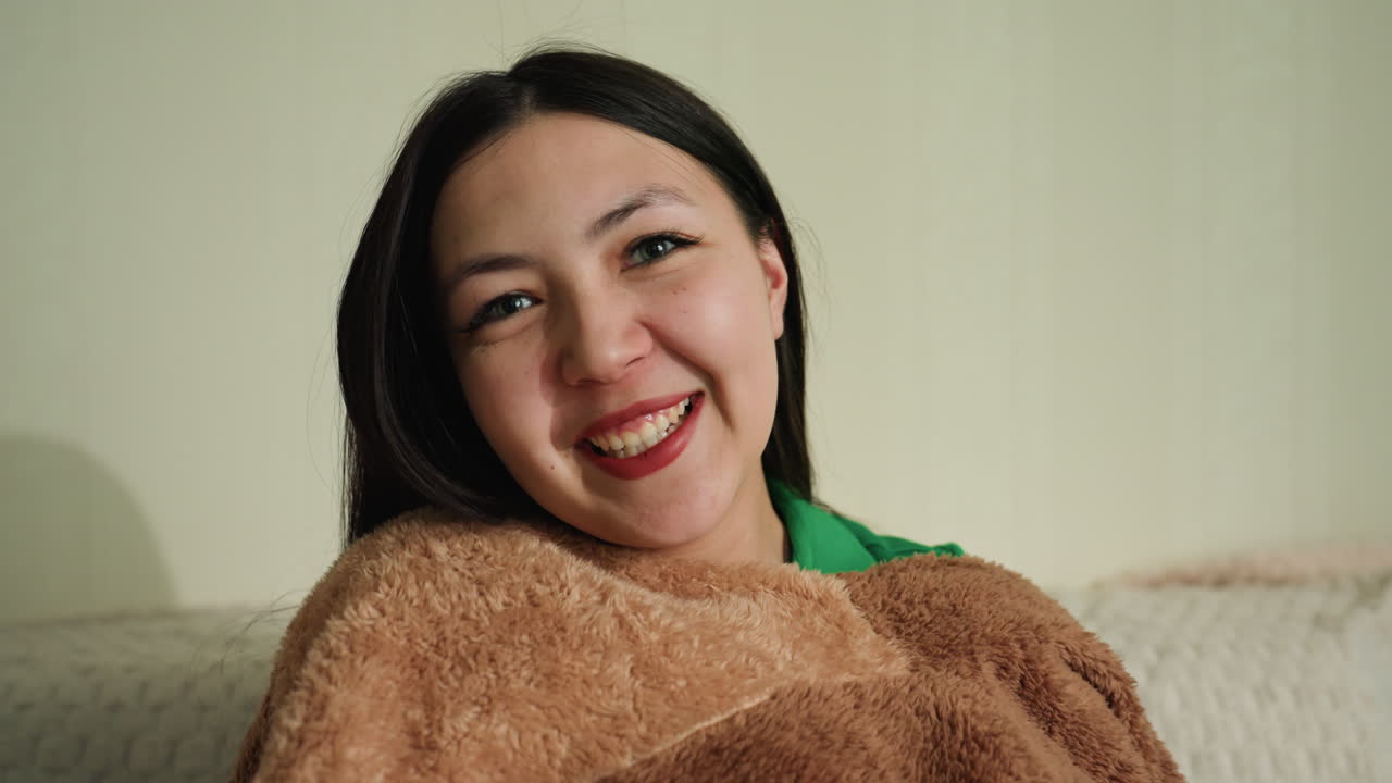 포근하게 미소 짓는 여성, 따뜻한 담요를 덮은 여성 클로즈업, 편안한 집에서의 순간을 즐기는 여성의 평온한 모습, 가정 환경에서 평온함과 따뜻함을 보여주는 부드러운 여성의 모습