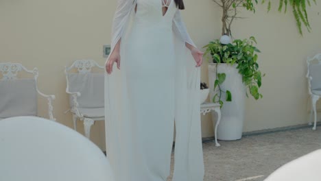 elegant bride in flowing white gown, softly holding her dress, standing in a serene garden