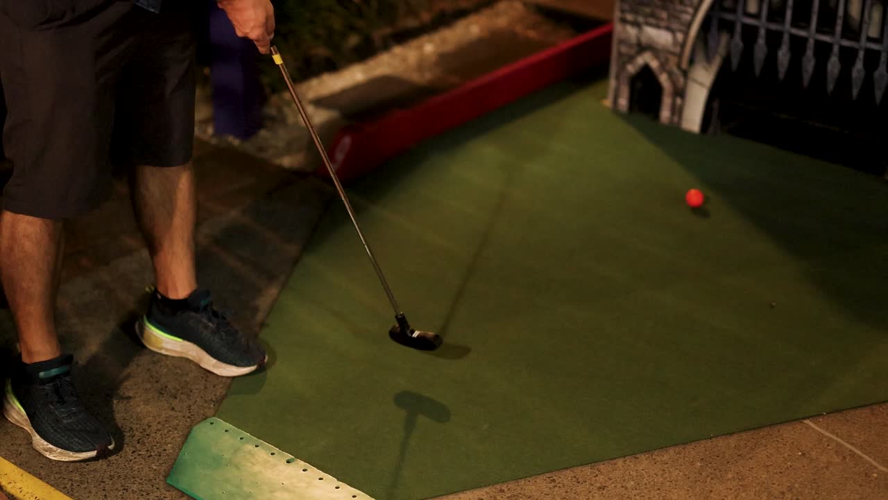 A person putts a golf ball on a mini-golf course at night, illuminated by artificial lighting
