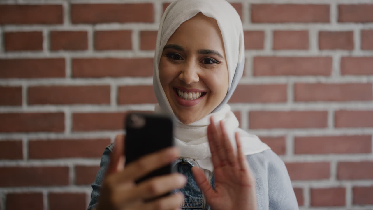 retrato de una joven musulmana feliz usando un teléfono inteligente video chat agitando la mano disfrutando de la comunicación móvil hermosa estudiante que lleva un pañuelo de hijab