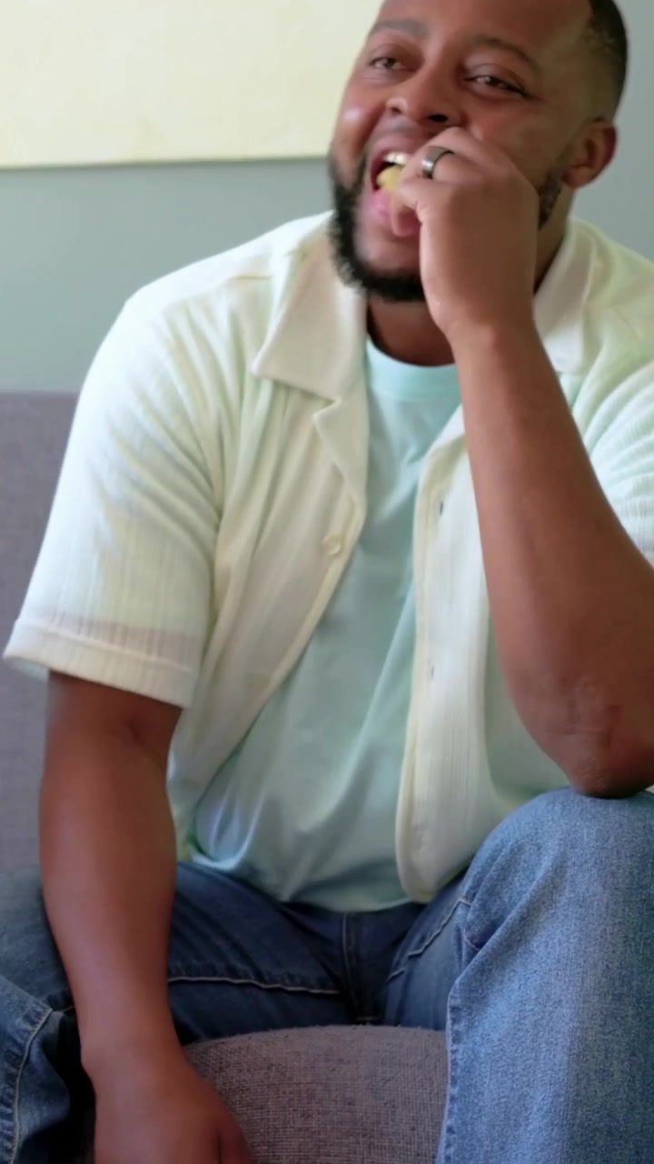 Video verticale: Sentendo un segnale fuori campo, uomo in camicia e jeans che tiene un gamepad e mangia una patatina sul divano.