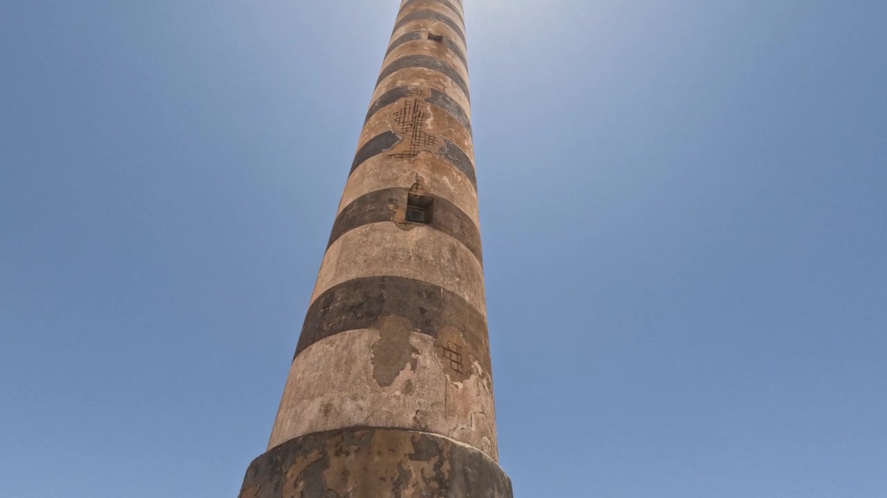 Storico faro alto a strisce che si erge in un paesaggio costiero arido, circondato da terreno sabbioso, muri consumati, vecchi edifici, vegetazione rada e un cielo azzurro limpido in un ambiente remoto