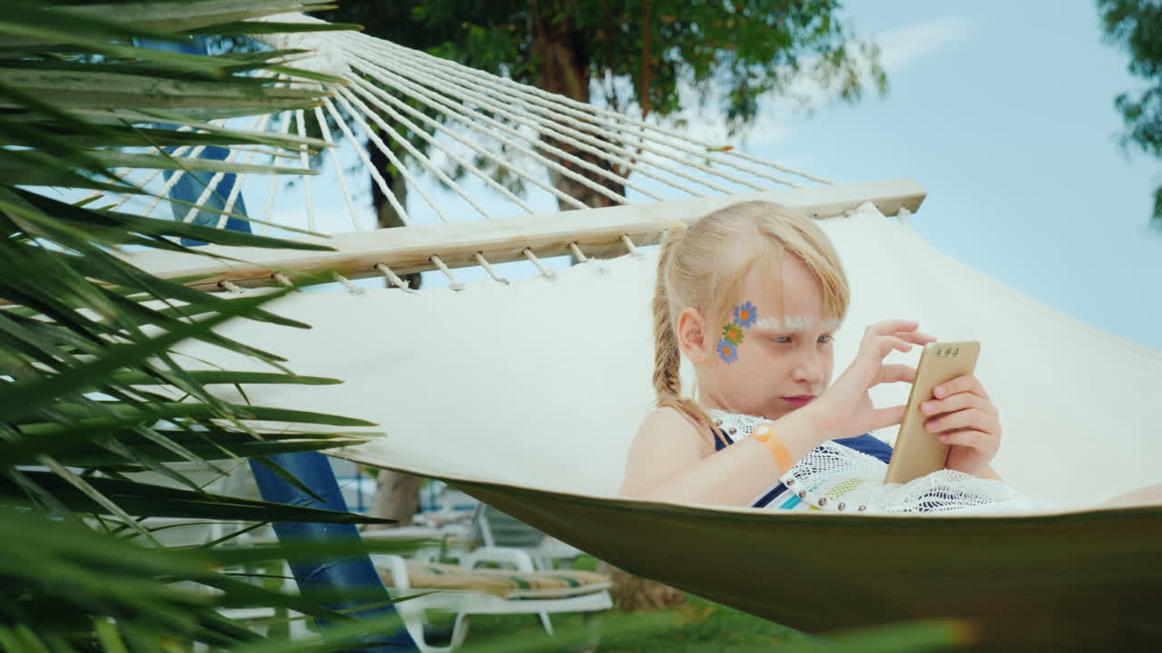 una chica genial con aquagrim en la cara está jugando por teléfono en una hamaca en un hotel turístico