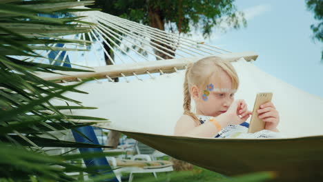 A-Cool-Girl-With-Aquagrim-On-Her-Face-Is-Playing-On-The-Phone-In-A-Hammock-In-A-Tourist-Hotel