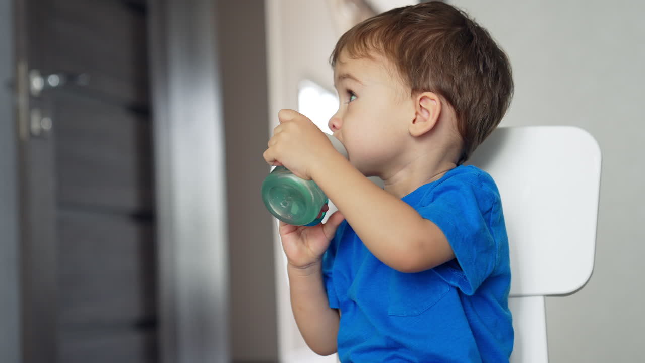 Adorable child drinking water from a bottle. Little kid sits on the chair and looks attentively aside. Blurred backdrop.
