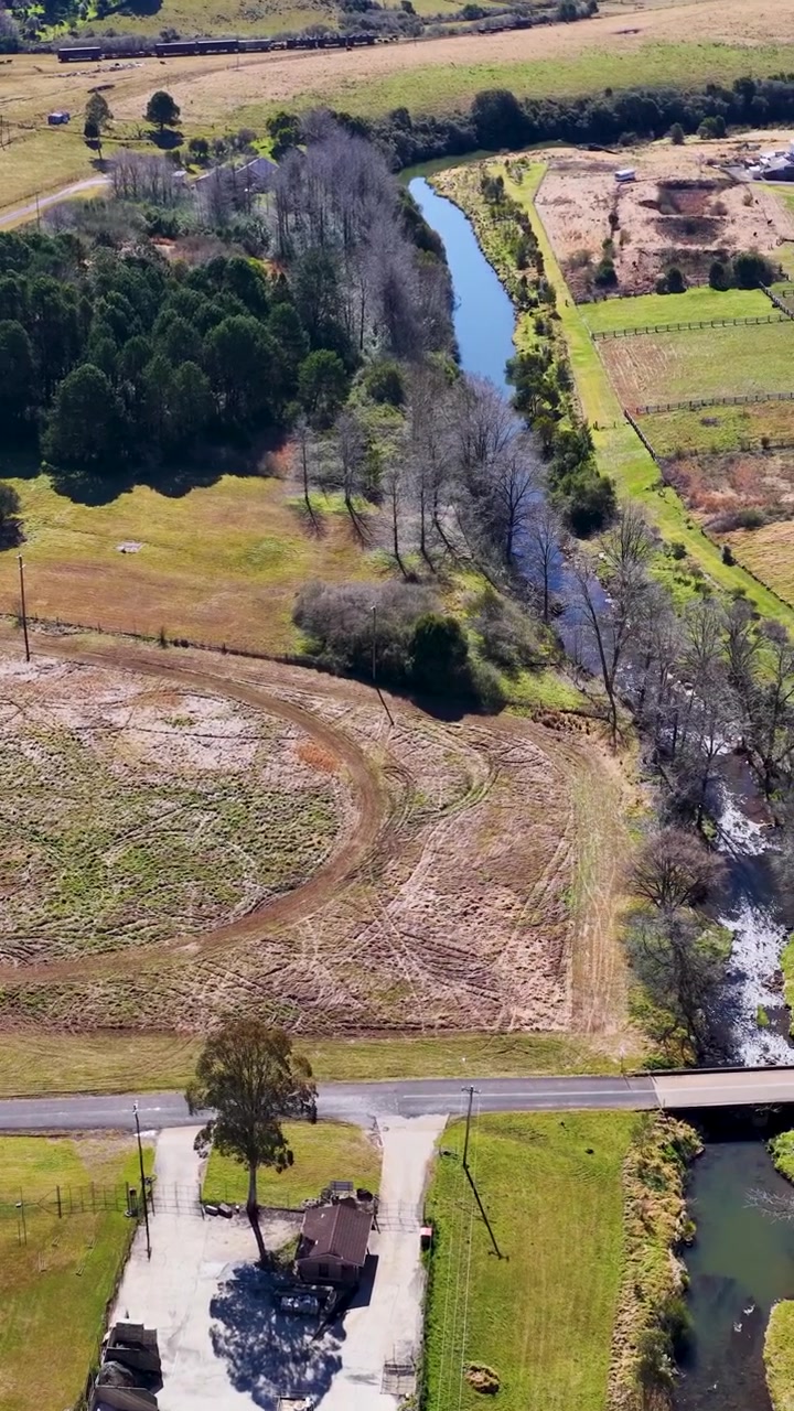 Dron Asciende sobre las Tierras de Cultivo de Dorrigo, Revelando Arroyo, Campos, Edificios Rurales y Luz Diurna Clara