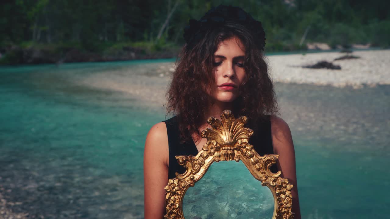 Dreamy Cinemagraph - seamless video loop of a beautiful young model girl standing at a fresh blue mountain river with an old golden mirror in Germany-Bavaria in a white fashion dress.