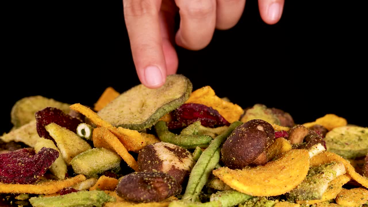 A hand picks assorted dried vegetable chips, including beetroot, potato, mushroom, and green beans, from a vibrant snack pile against a black background under bright studio lighting