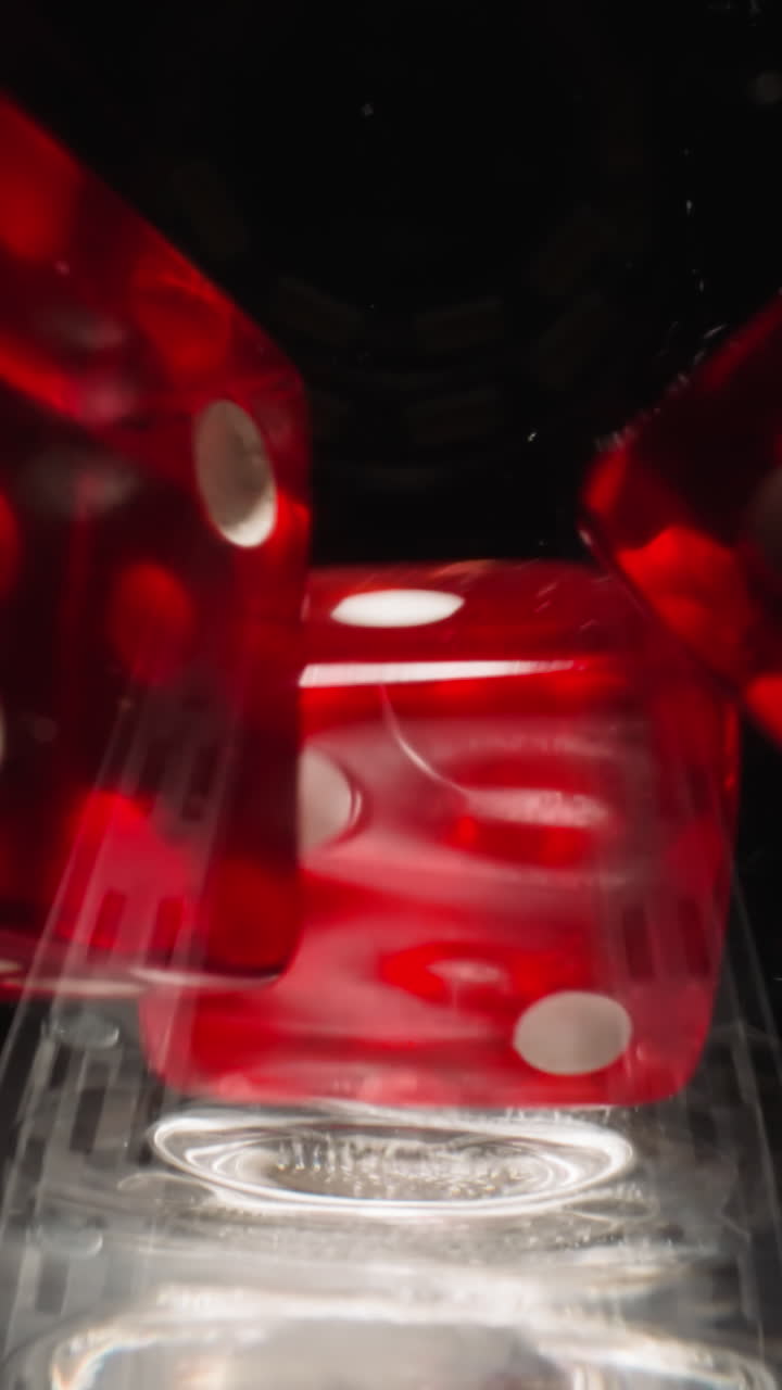 Red cubes with white dots falling into wineglass spin at bright light in studio. Dice for games hitting to sides of glass with fine reflection stop on black background macro
