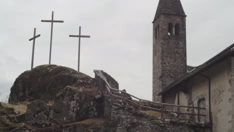 camera tilts up towards a small rock church with three crosses alongside, in a wood of the italian alps 4k slow motion bell tower detail