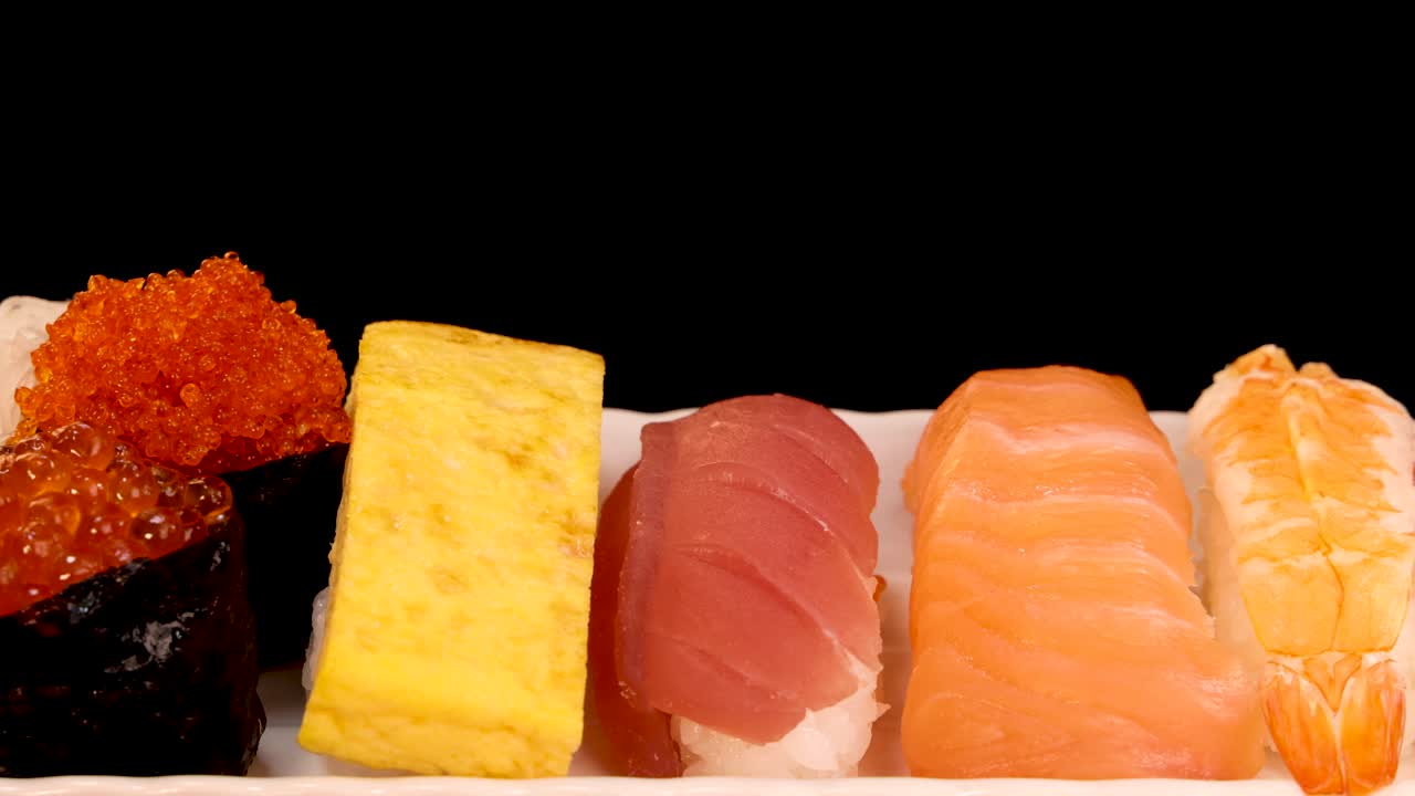 A white plate of assorted sushi, including nigiri, sashimi, and tamago, rotates smoothly against a black background under bright, even lighting