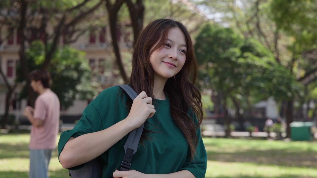晴れた大学のキャンパス公園で、若い女性が微笑みながらバックパックを背負っている。