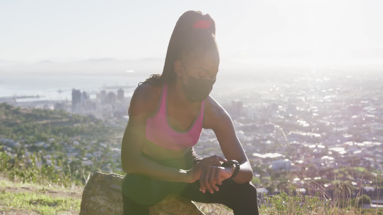 mujer afroamericana que usa una máscara facial usando un reloj inteligente mientras está sentada en una roca al aire libre