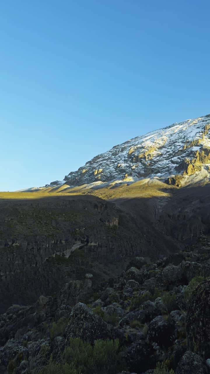 Statisk billede i jordhøjde viser en klippekant med de sneklædte skråninger af Mount Kilimanjaro i baggrunden under en klar, blå himmel