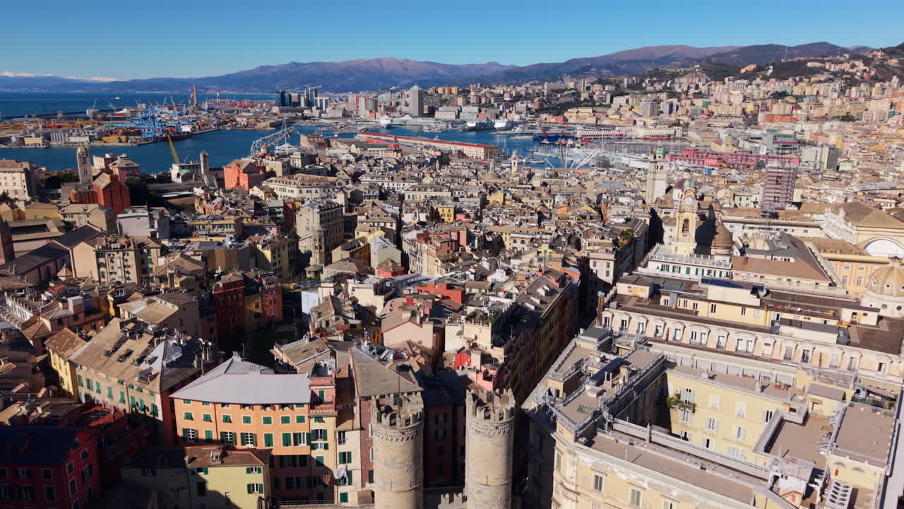Un drone vola sopra le torri di Porta Soprana a Genova, Italia, muovendosi sul denso centro storico verso il porto antico, il porto commerciale e le montagne liguri sotto un cielo sereno.