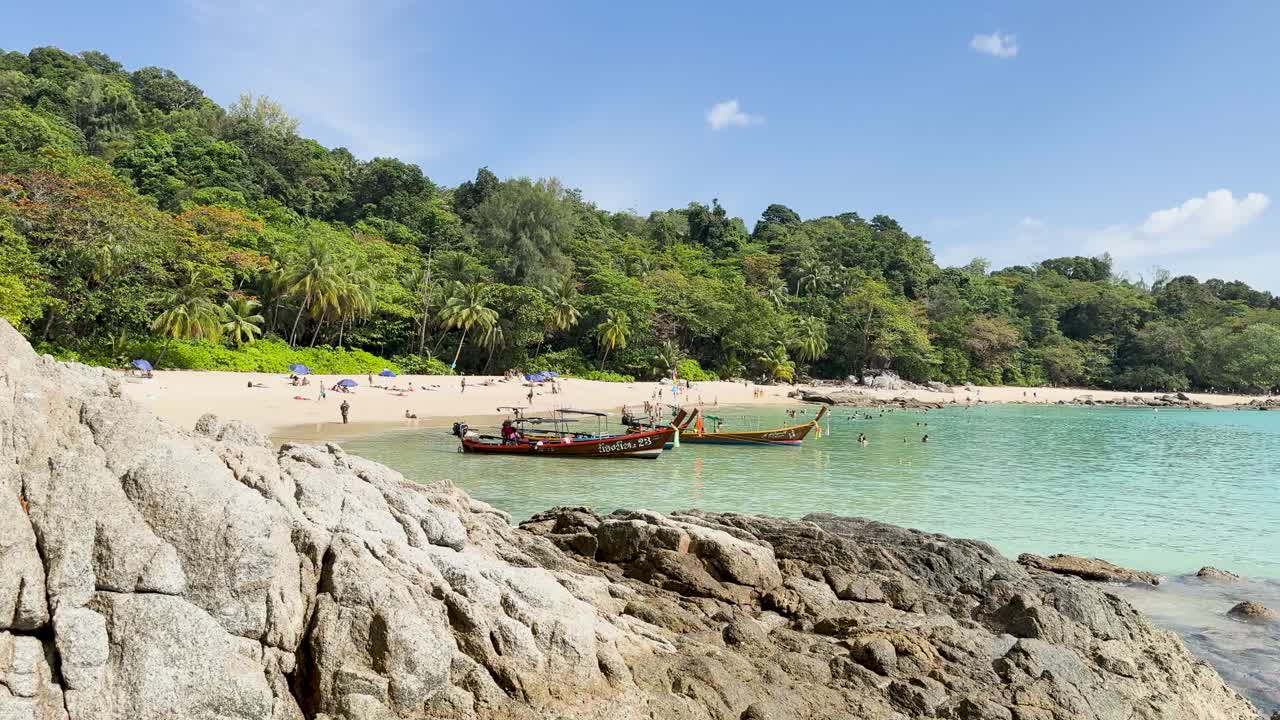 A tranquil beach in Phuket with clear waters, lush greenery, and traditional boats under bright daylight