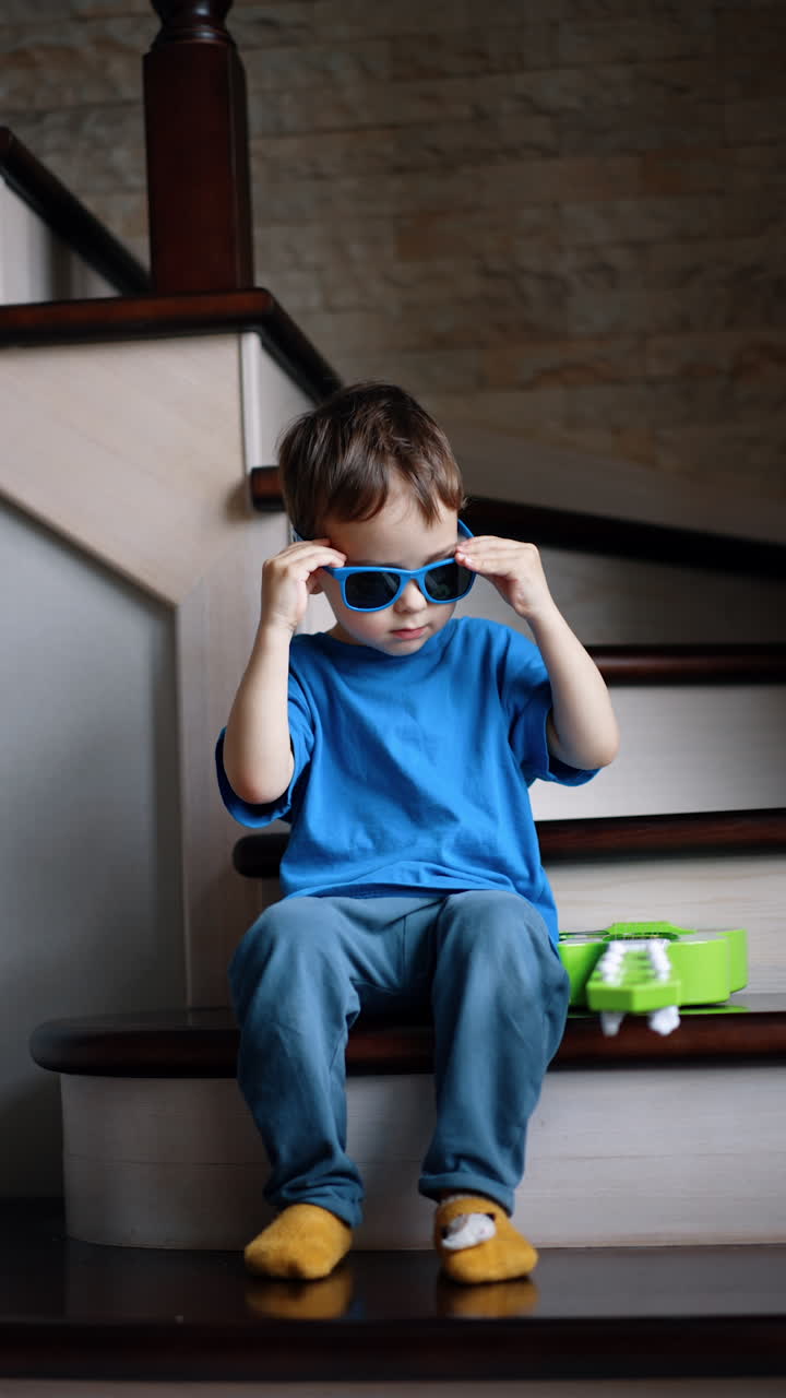 Serious cute kid sitting on the stairs puts off his little green guitar. Toddler takes off his sunglasses and puts near the instrument. Vertical video.