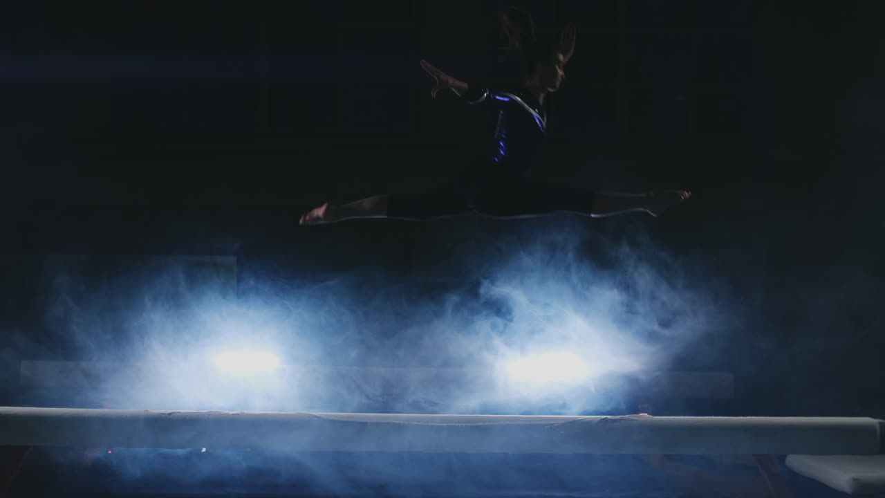 una chica en un traje de baño deportivo para gimnasia hace un salto en cámara lenta mientras está de pie en una viga de equilibrio