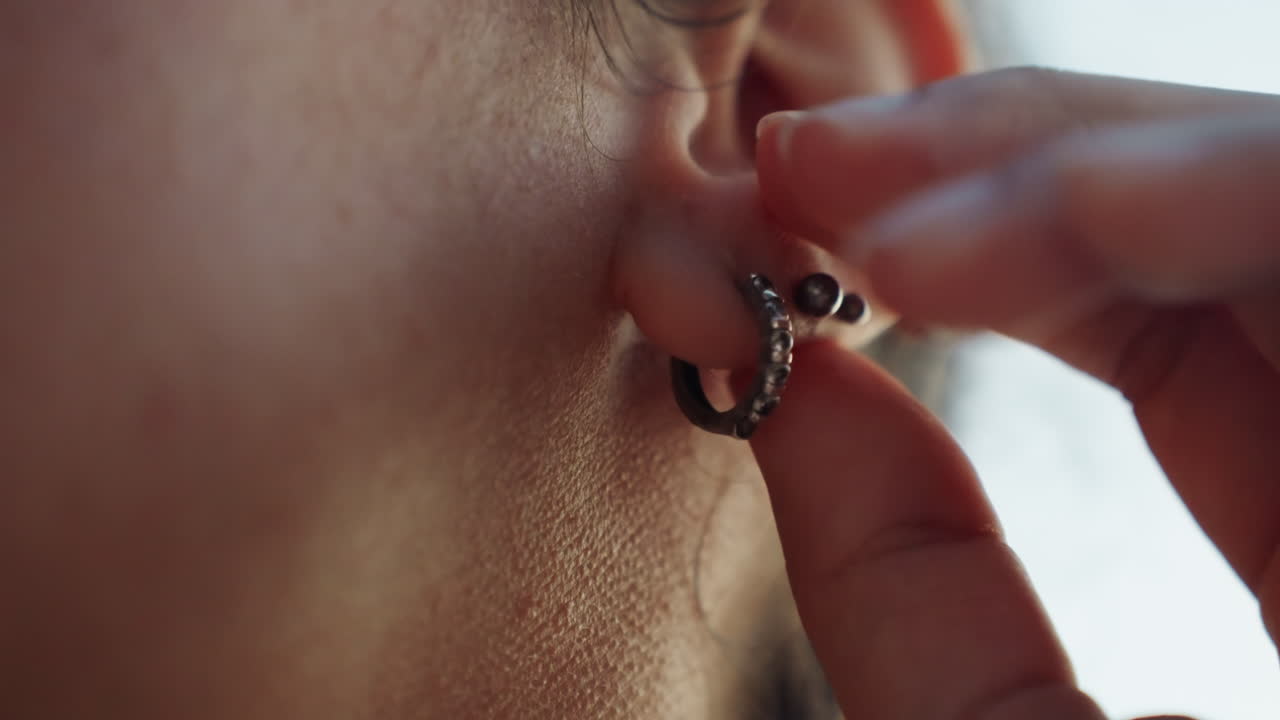 Primer plano de oreja con pendientes tocándose, artesano ajustando un aro de plata, suave yema del dedo, luz natural suave, textura de la piel y detalle del lóbulo visibles, estilo delicado y rutina matutina tranquila, poca profundidad de campo.