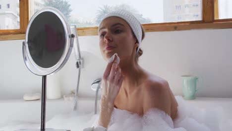 woman cleansing her face with cotton pad while sitting in bathtub