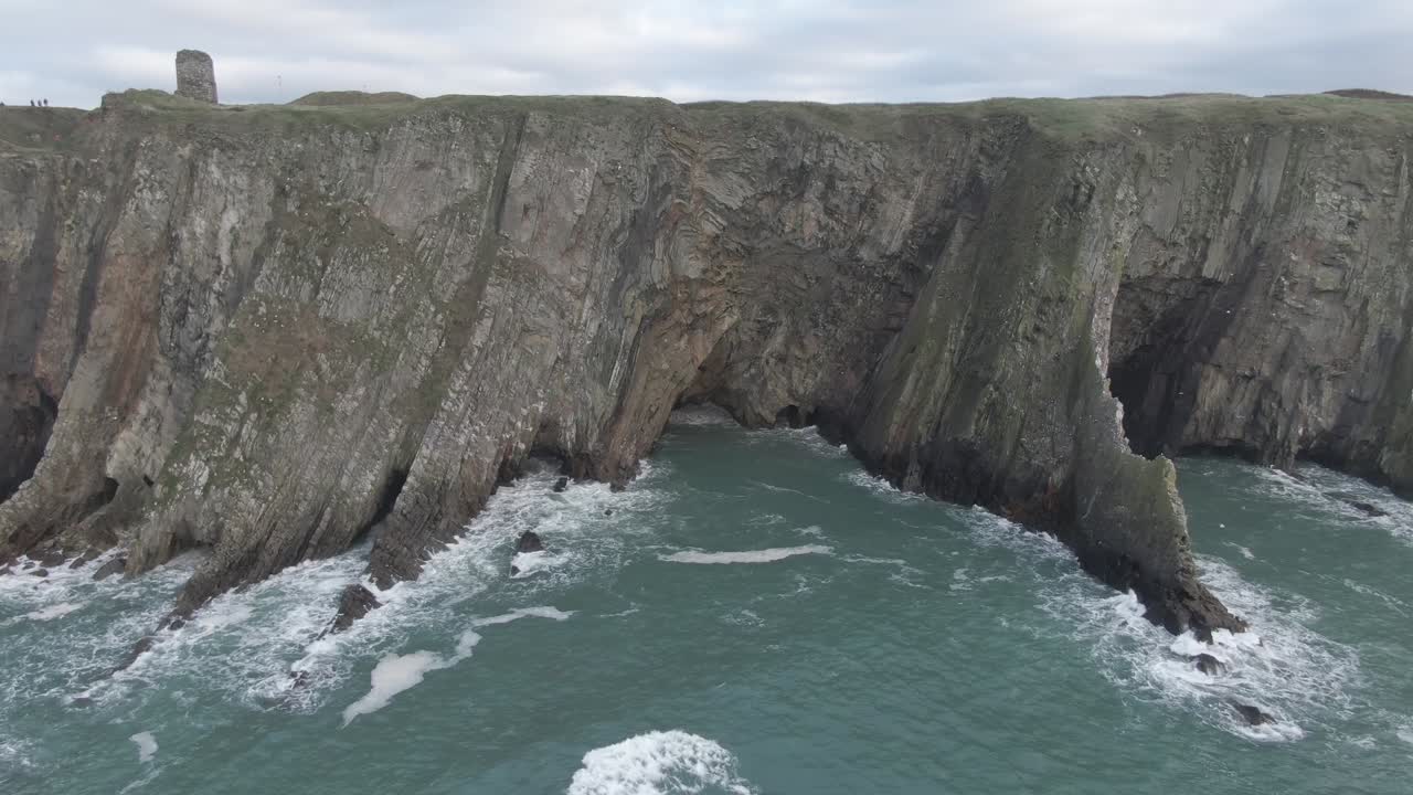 Stationary aerial view of sea side cliffs with rough seas and waves hitting the cliffs, steep cliffs and castle ruins