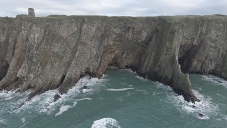 stationary aerial view of sea side cliffs with rough seas and waves hitting the cliffs, steep cliffs and castle ruins