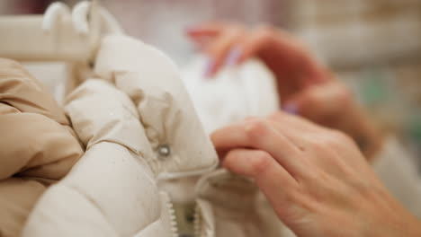 hand view of woman checking neck of winter coat in cloth store, fingers exploring padded quilted collar and knitted cuffs, inspecting textile quality, pastel jackets blurred in background