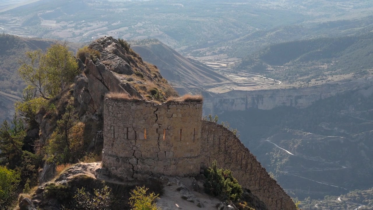 Rovine di una fortezza erosa coronano un ripido crinale montuoso, affacciandosi su una vasta valle nebbiosa, strade pallide e tortuose, scogliere aspre, arbusti sparsi ed erba secca sotto una luce diurna soffusa, creando un'atmosfera remota.