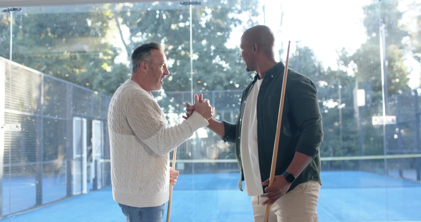 Diverse friends greeting after man entering right, gathering near glass wall holding cues playing