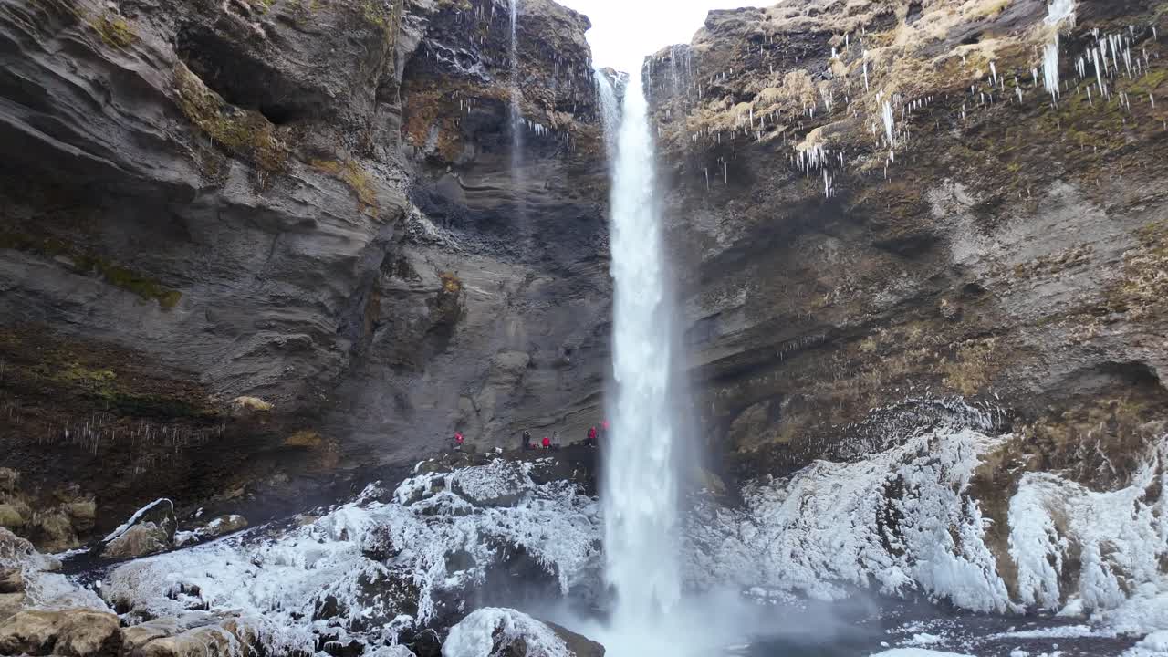 The video showcases the serene Kvernufoss waterfall in Iceland, nestled in a lush green valley, with water flowing gracefully down a cliff, surrounded by dramatic cliffs and tranquil nature.
