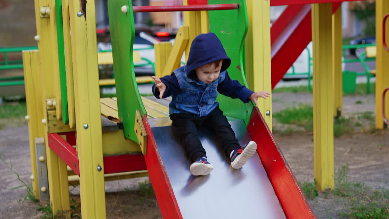 Baby boy sits on the slide tapping his feet. Kid in jeans jacket goes down and runs to the ladder.