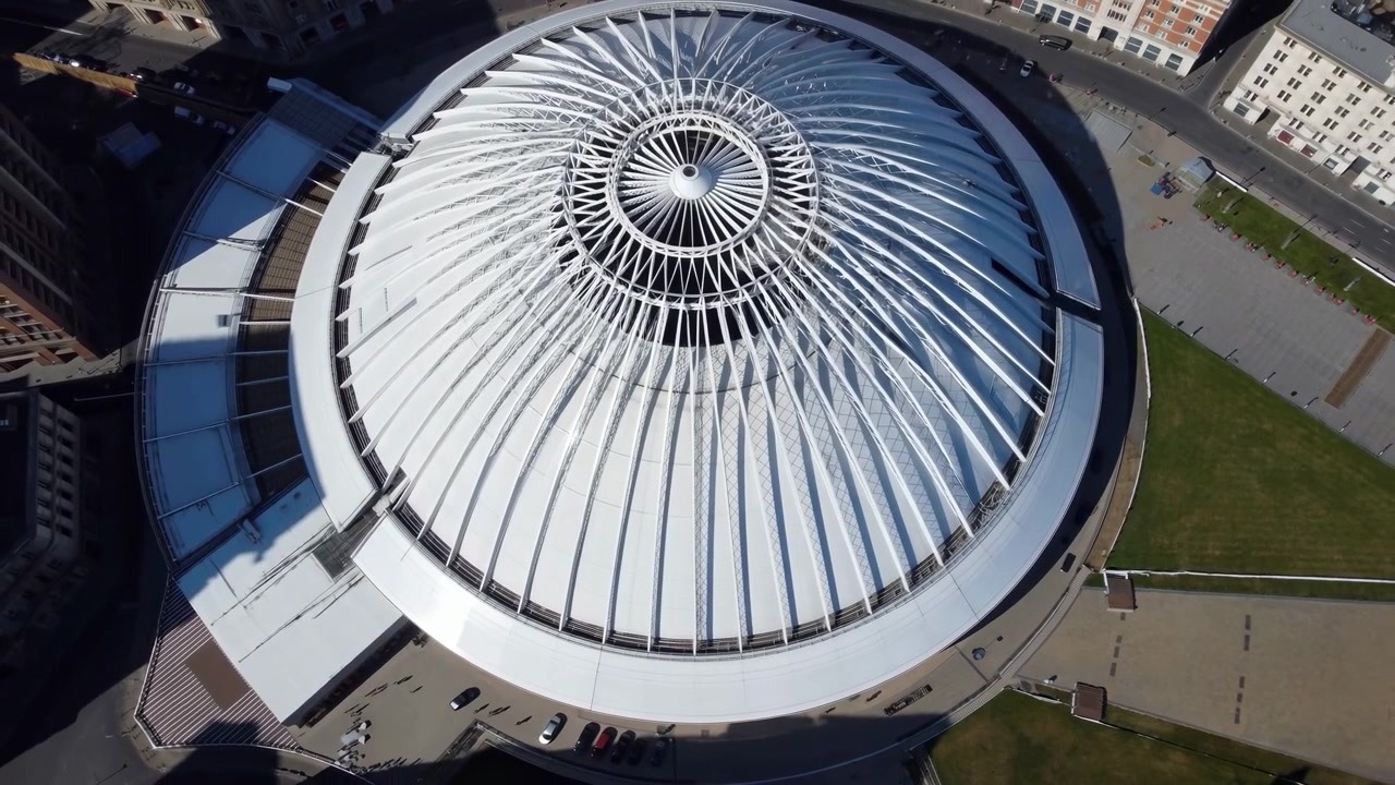 Vista dall'alto di un grande edificio pubblico rotondo con un tetto a coste luminoso, lucernario centrale, passerelle circostanti, aree verdi e forti ombre diurne