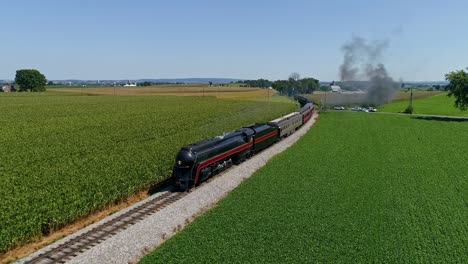una vista aérea de un tren de pasajeros de vapor antiguo que se acerca después de dar la vuelta a una curva que sopla humo y vapor que viaja a través de campos de maíz fértiles durante la hora dorada en un día soleado de verano